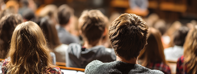 Rows of students sit in a lecture hall. The view is from the backs of their heads.