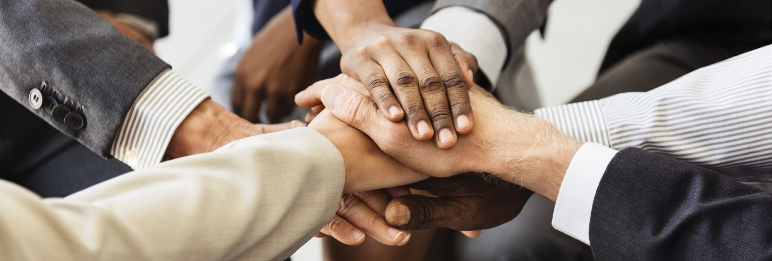 Closeup of several individuals’ hands clasped over one another, as in a team huddle. Individuals are all wearing formal business attire.”