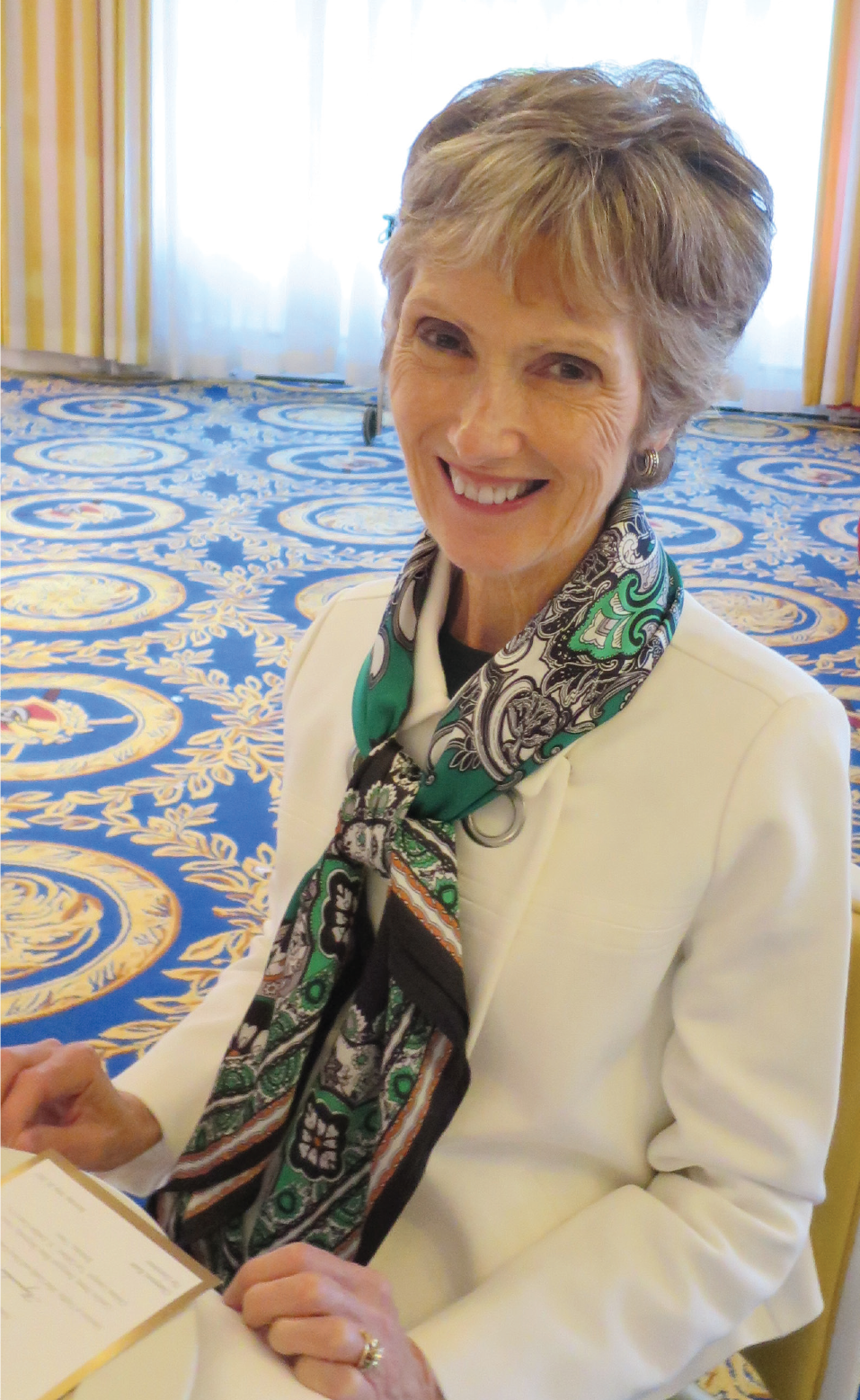 Photo of Diane Bosse sitting at a table, smiling, wearing a white suit jacket and green scarf