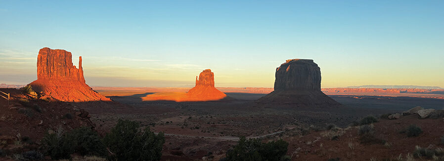 A photo of Monument Valley Navajo Tribal Park. Three rocky sky islands appear in the far-away distance; one is cast in shadow, and the other two glow bright orange in the rising sun. The surrounding desert landscape is also cast in shadow. The sky is a gradient of light yellows and blues.