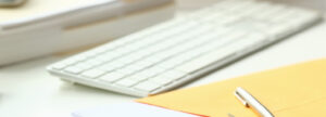 Image of a wireless white keyboard on a desk, with a manila folder and pen resting in front of it