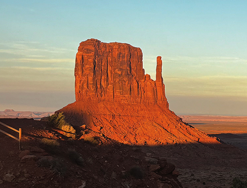 A photo of Monument Valley Navajo Tribal Park. Three rocky sky islands appear in the far-away distance; one is cast in shadow, and the other two glow bright orange in the rising sun. The surrounding desert landscape is also cast in shadow. The sky is a gradient of light yellows and blues.