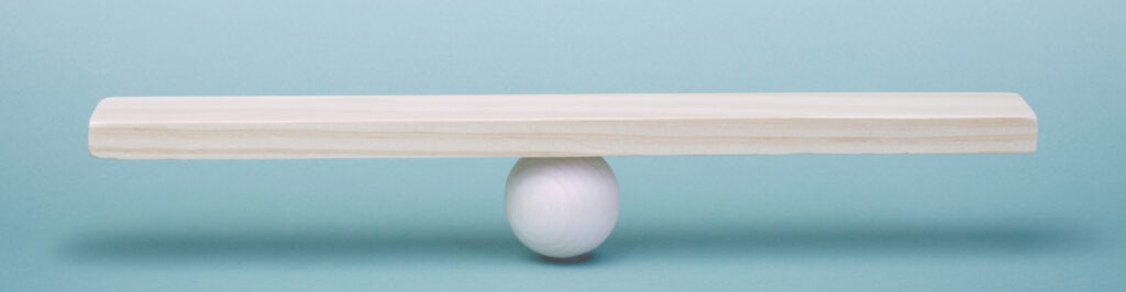 On a blue background, a long pale wooden board is centered and balanced on a white wooden ball