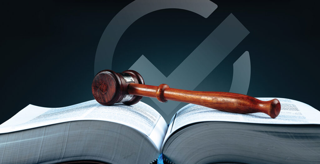 A wooden gavel lays across an opened textbook. The backdrop is solid black with a gray check mark.