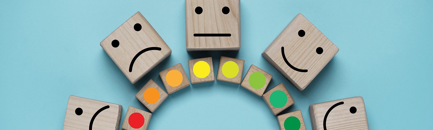 an arc of wood blocks with color dots ranging from deep red on the left, through orange and yellow, to green on the right. Four larger wood blocks are arranged above the arc, with simple faces drawn on them. The faces range from a deep frown on the left to a big smile on the right.”