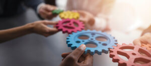 Photo of several hands holding gears of various colors horizontally over a table so that the gears interlock