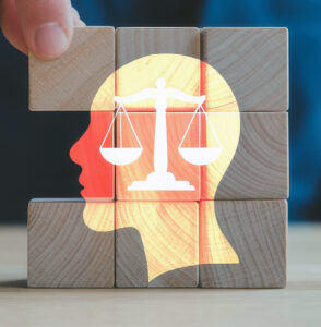 Wooden blocks are stacked in a 3-by-3 layout with one missing in the center. Printed on the blocks is a head silhouette, and in the center are the scales of justice. The backdrop is a gradient of blueish gray.